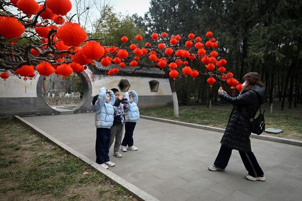 Група деце позира за слику испред црвених фењера окачених на дрвеће за прославу кинеске лунарне Нове године у парку у Пекингу 11. јануара 2023. - Sputnik Србија