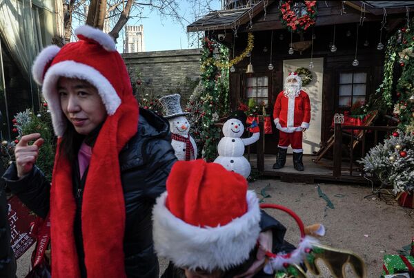 Жена и дете носе свечане капе док пролазе поред радника обученог у одело Деда Мраза који чека посетиоце на божићном вашару и пијаци са француском тематиком  у Пекингу. - Sputnik Србија