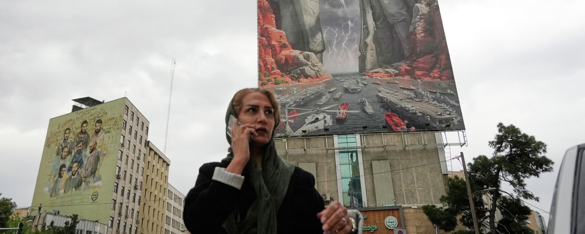 Ženщina c mobilьnыm telefonom na fone plakata s izobraženiem iranskogo voenačalьnika Raisa-Ali Delьvari i komanduющego VMS KSIR Alirezы Tangsiri v Tegerane, Iran - Sputnik Srbija, 1920, 23.04.2026