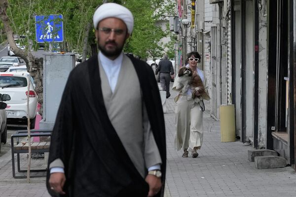 Жена носи свог пса у шетњи  иза имама који хода тротоаром у Техерану. - Sputnik Србија