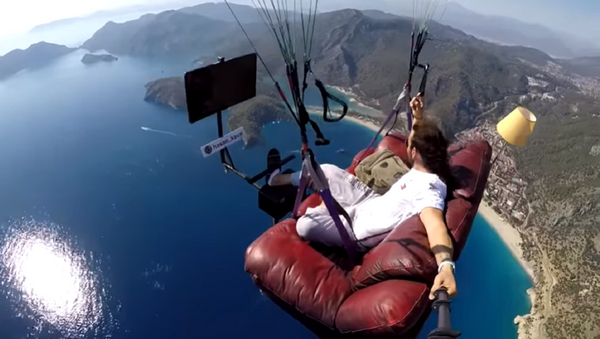 A screenshot of a video of Hasan Kaval, a Turkish paragliding instructor, watching TV while flying over water on a couch attached to a paraglider. - Sputnik Србија